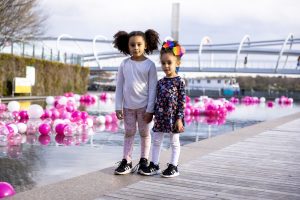 Two Cherry Blossom fans pose in front of outdoor art installations for the 2022 Petalpalooza. The Yards Park Bridge behind them features the artwork of Park West favorite Daniel Wall (Credit: National Cherry Blossom Festival)